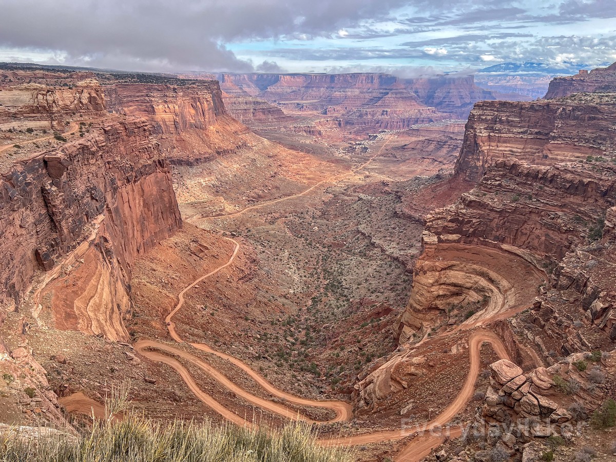 The windy, hairpin turns of Shafer Trail road descending 1,400 ft to the canyon floor below. Steep cliff walls may be seen on both sides of the frame.