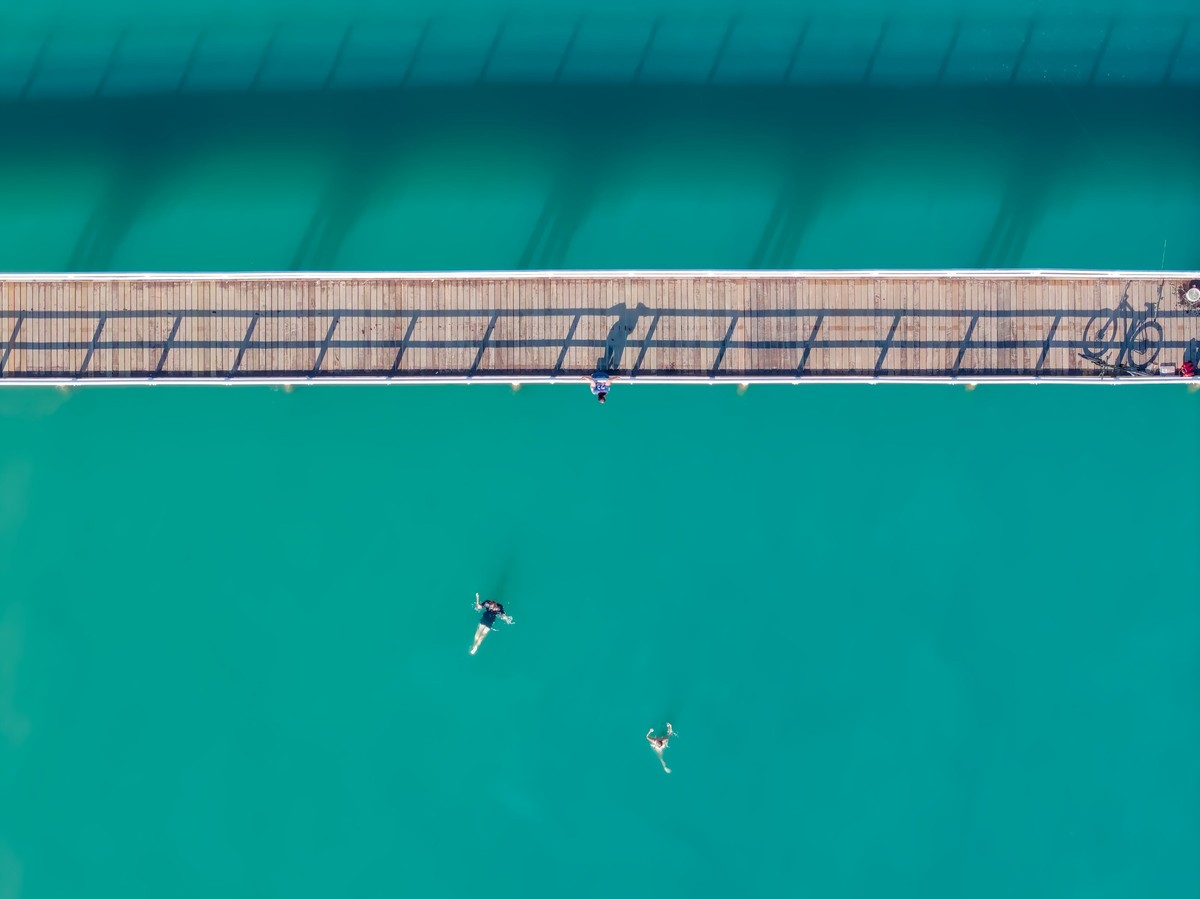 A top down shot of a narrow wooden pier, sitting in aqua blue water.  Two kids are visible swimming in the water looking up at the pier, where a 3rd kid hangs off the outside of the rails, trying to build the courage to jump
