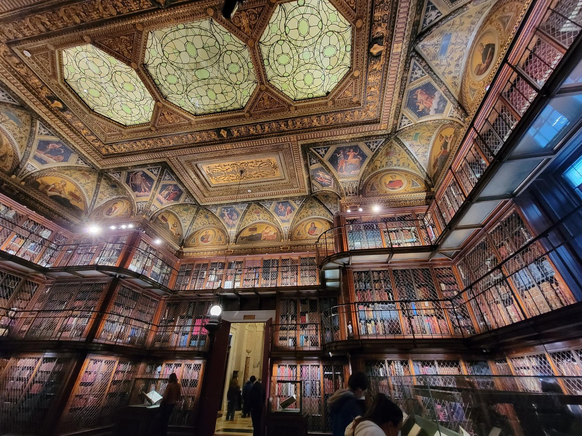 wide-angle view of elegantly-lit exhibit room with three levels of bookcases covering the entire wall. the edges of the ceiling are made of vaulted panels with classical scenes, and the center is an art-deco skylight with light green interlocking oval designs. on the ground are people looking at exhibits.