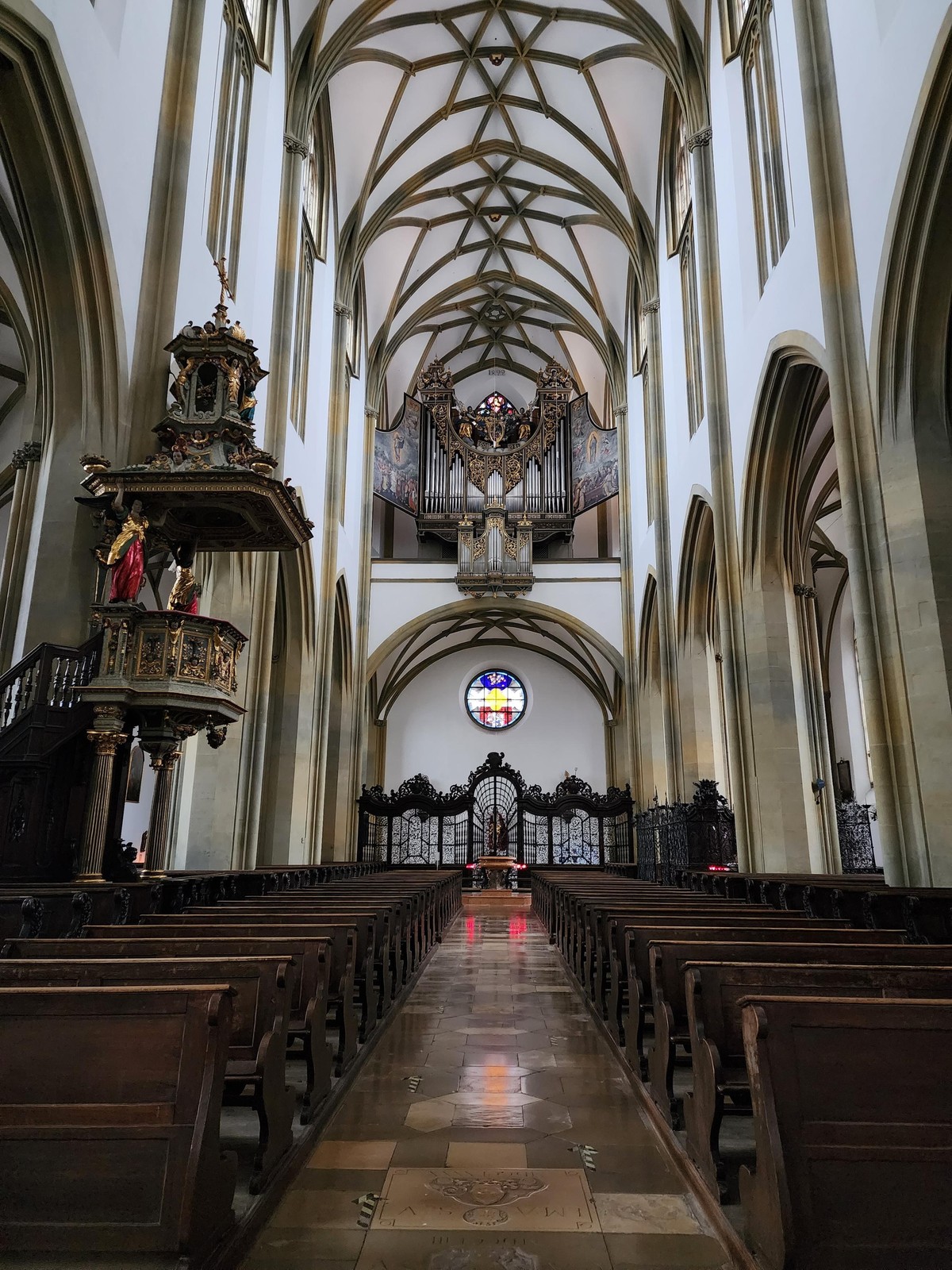 Looking from the altar towards the organ, which is high overhead and has open panels with religious scenes that cover the pipes when closed. Below it is a roundel window. The walls and ceiling are the familiar white with prominent brown ribbing/vaulting, and there are two rows of pews running from the foreground almost to the (organ) wall.