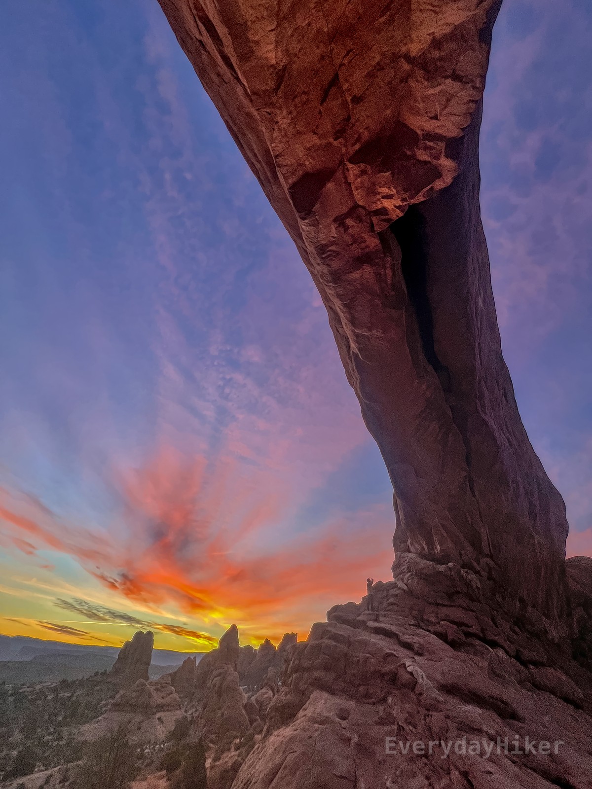 The large bridge that makes North Window passes overhead while the left half of the frame is bathed in vibrant sunrise yellows and oranges. A person looking up at the arch from the other side helps understand the scale of it.
