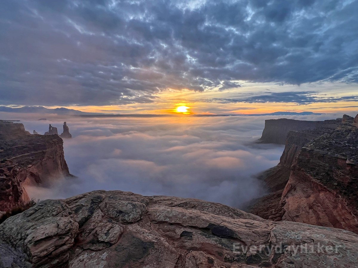 Receding fog reveals more of the steep cliffs along this horseshoe, with a few tall and skinny spires to the left in front of a distant mountain range. The rising sun sits in between the fog and the clouds in the center of frame.