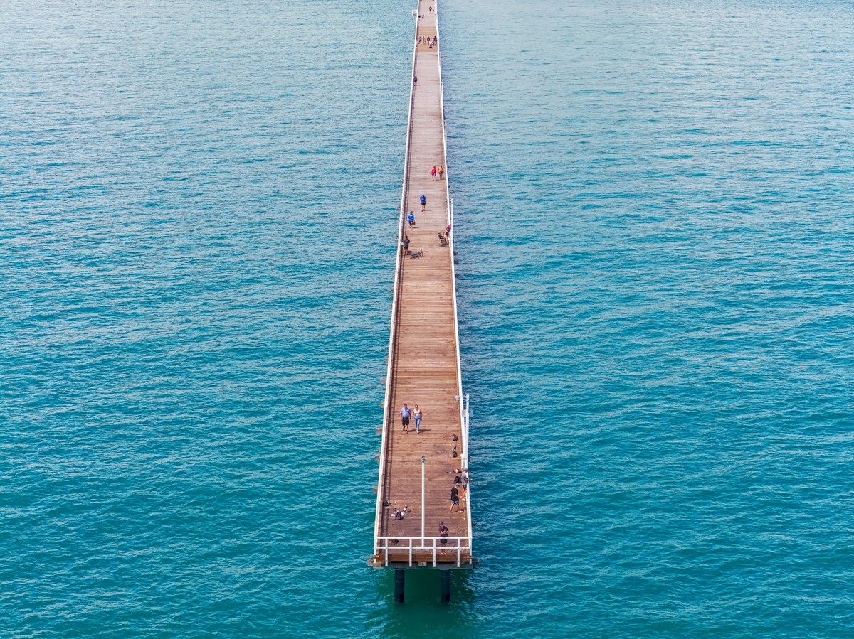 A drone photo taken from the ocean, looking towards the short, back along the length of a very long pier. The camera is angled down slightly however, and the shore is not visible due to the framing.