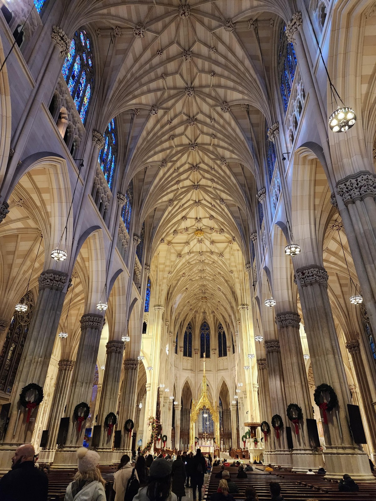 View of the altar from the entrance, showing much of the length of the ribbed vaulted ceiling and stained glass windows above the nave. in the distance is the apse with the altar under a gold canopy or altarpiece. on either side are columns dividing the main nave from the side naves, and there are people filling the aisle and pews.
