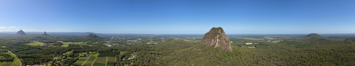 A panoramic drone photo showing the tree covered plains of the Glass House Mountains area, with the Glass House mountains themselves popping out here and there out of the landscape